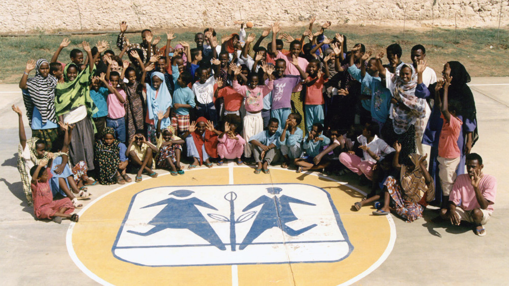Eine große Gruppe von Kindern und Erwachsenen stehen und sitzen um ein SOS-Kinderdorf-Logo, das auf den Boden gemalt wurde
