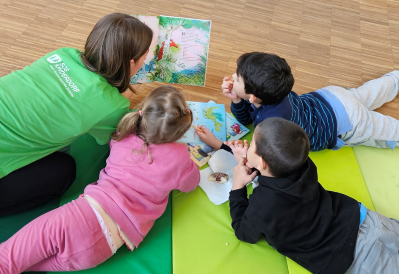 Runder Tisch mit Kinderbüchern im Vordergrund, lesende Kinder auf gemütlichen Matten am Boden im Hintergrund.