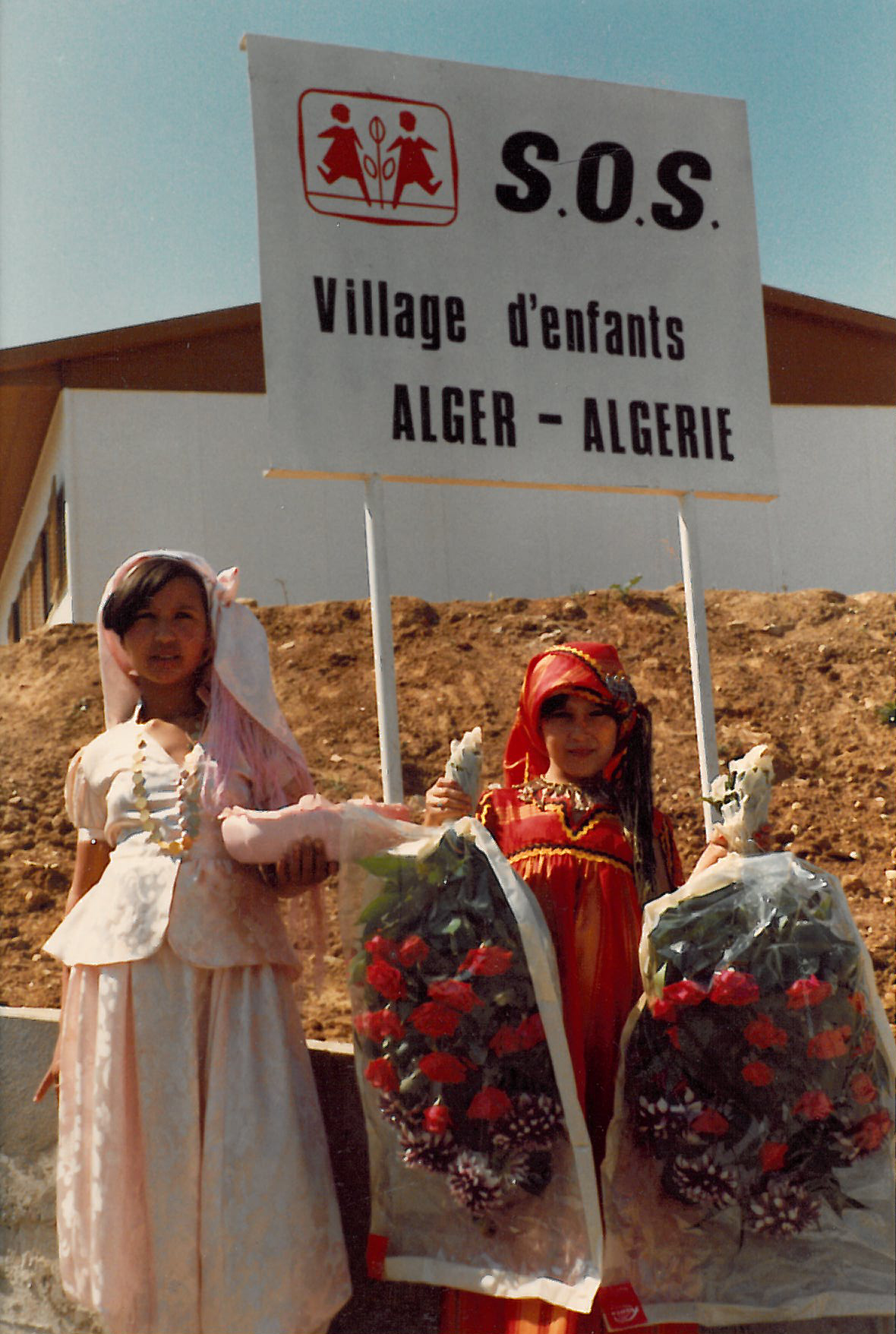 Zwei Mädchen in traditionellen Gewändern und mit Blumen vor einem Schild mit der Aufschrift "SOS Village d