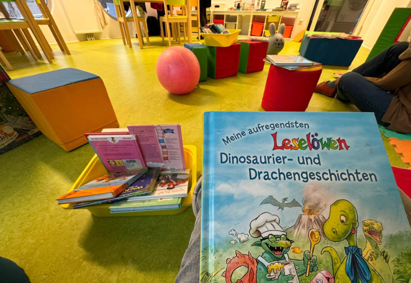 Kinderbuch "Meine aufregendsten Dinosaurier- und Drachengeschichten" im Vordergrund, kindergerechte Einrichtung mit Sitzgelegenheiten und einem Behälter mit weiteren Kinderbüchern im Hintergrund