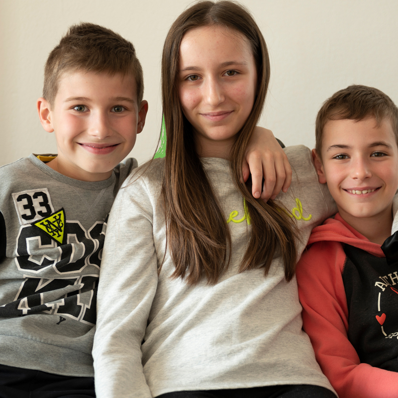 eine frau mit hellbraunen, langen haaren und brille sitzt mit einem kind auf dem schoß beim küchentisch und blickt zum herrn, der am linken bildrand verschwommen zu sehen ist