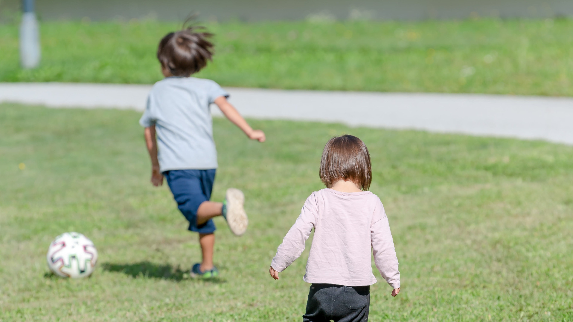 Zwei Kinder mit dem Rücken zur Kamera fotografiert, das im Hintergrund läuft einem Ball hinterher.
