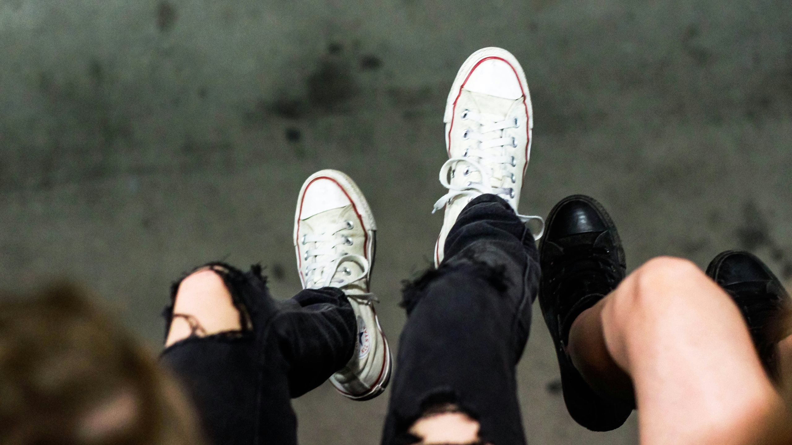 Blick von oben auf baumelnde Füße von jungen Erwachsenen. In der Mitte sind Beine in einer zerrissenen Jeans und mit weißen Converse Schuhen.