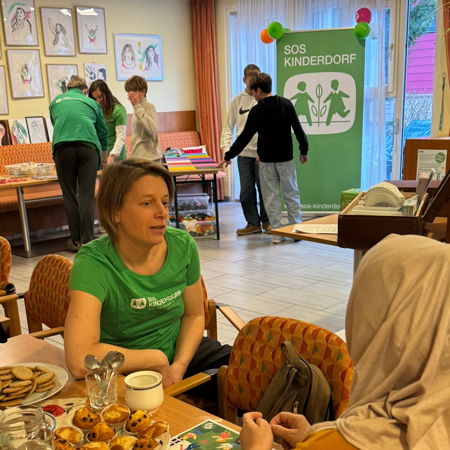 Momentaufnahme im Familiencafé, im Vordergrund zwei Erwachsene im Gespräch bei Kaffee und Muffins, im Hintergrund Kinder beim Legotisch