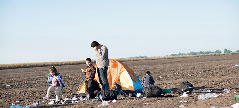 3 Flüchtlingskinder und ein Erwachsener stehen vor einem Zelt inmitten eines Feldes, umgeben von Müll 