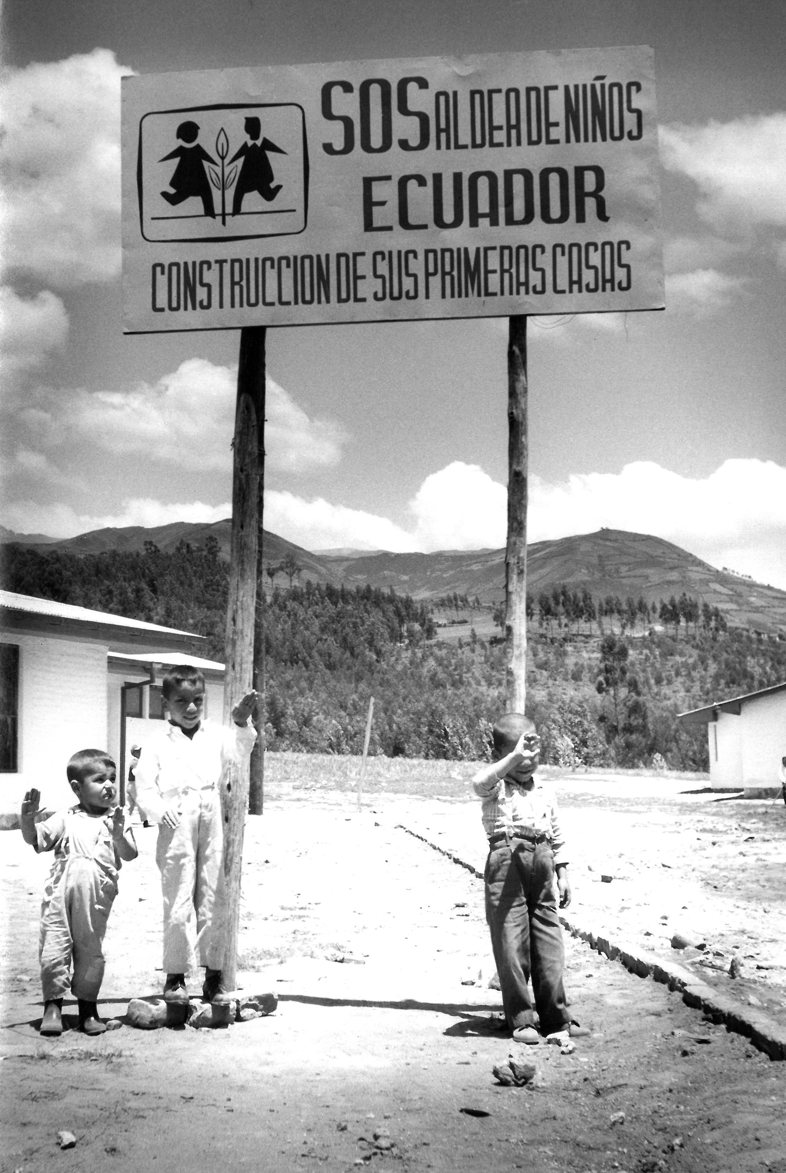 Drei Kinder stehen vor einem großen Schild mit der Aufschrift "SOS Aldea de Ninos Ecuador. Construccion de sus primeras casas"