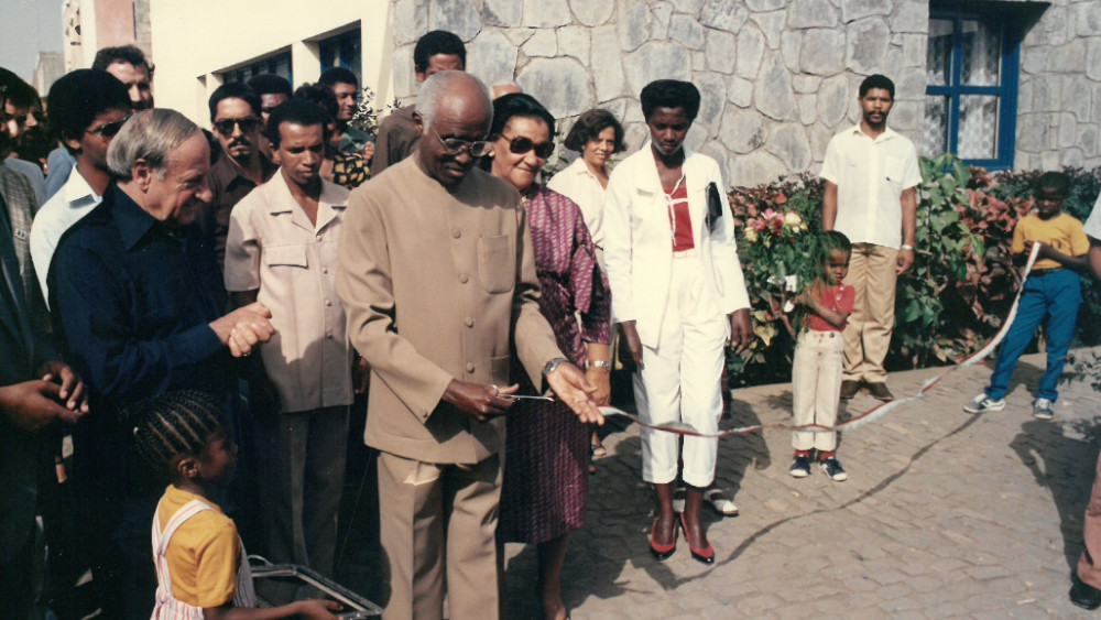 Einige Menschen in feierlicher Kleidung vor einem Absperrband, das ein Mann gerade durchschneidet