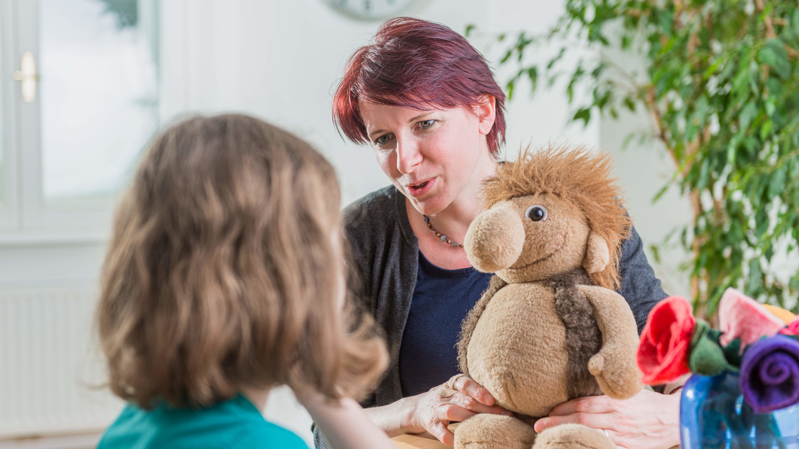 Foto von einer erwachsenen Person, einem Kind (nur der Hinterkopf im Vordergrund sichtbar) und einem Stofftier.