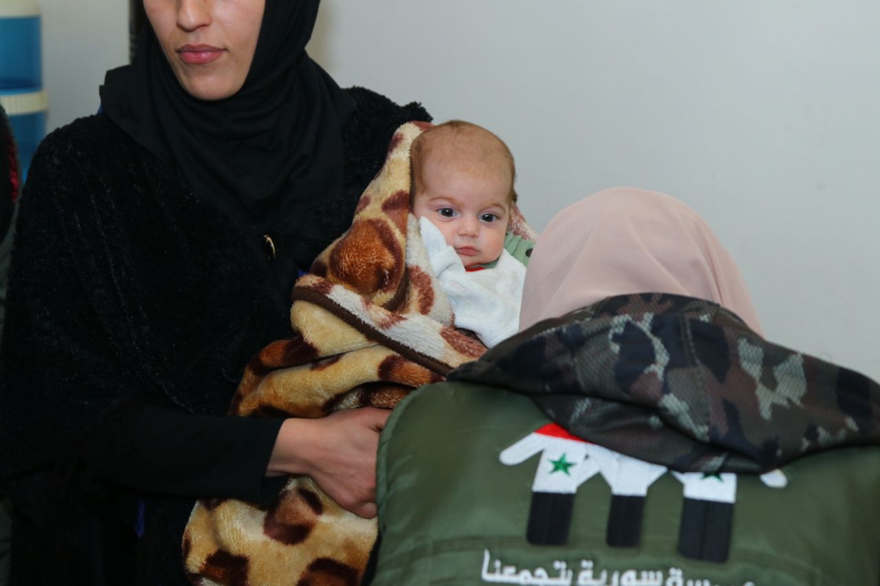 kleines Baby auf dem Arm einer Frau, im Vordergrund eine weitere Frau mit dem Rücken zur Kamera