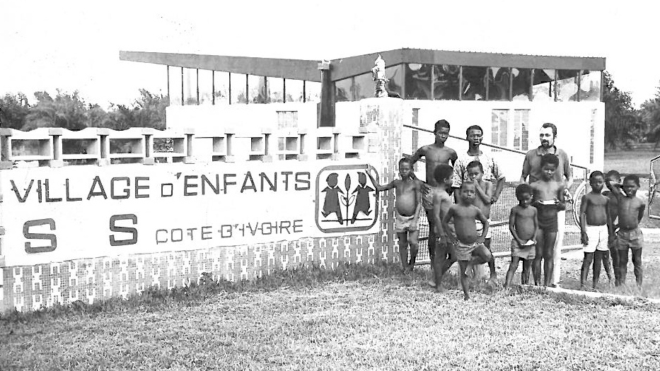 Einige Kinder und Erwachsene vor einem SOS-Kinderdorf, daneben die Tafel des Dorfs mit der Aufschrift "Village d