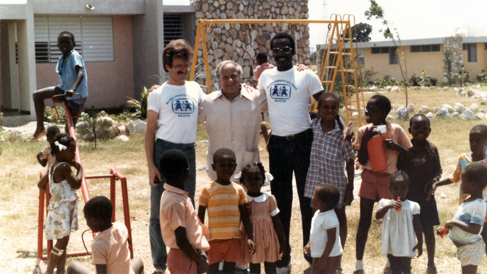 Hermann Gmeiner gemeinsam mit zwei Männern im SOS-Kinderdorf-T-Shirt umringt von einigen Kindern