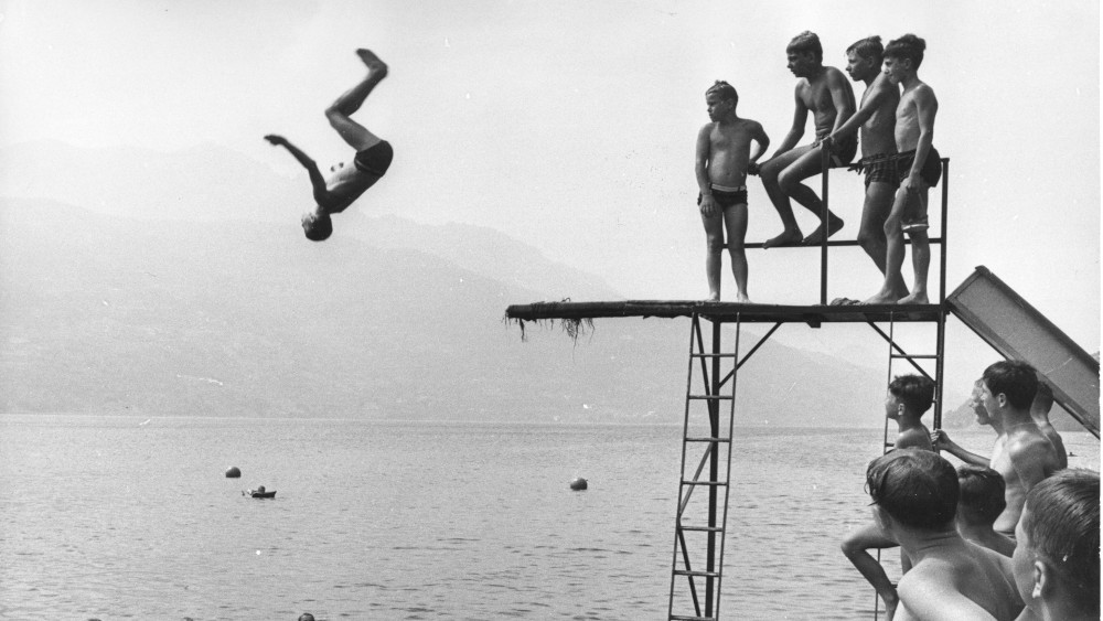 Junge macht einen Salto ins Wasser von einem Springturm, auf dem weitere Kinder stehen
