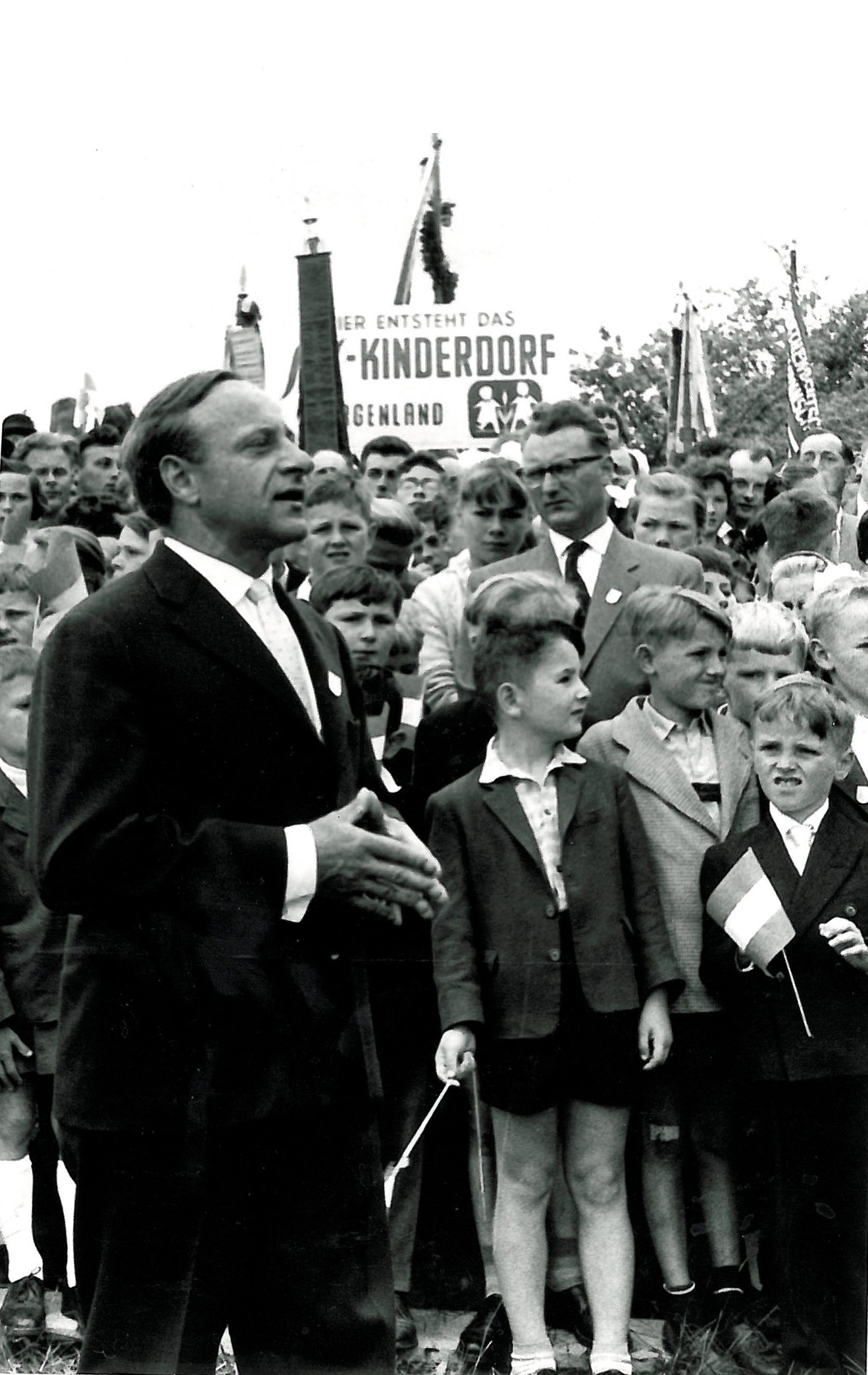 Hermann Gmeiner vor einer Menge an Menschen, im Hintergrund ein Transparent mit der Aufschrift "SOS-Kinderdorf Burgenland"