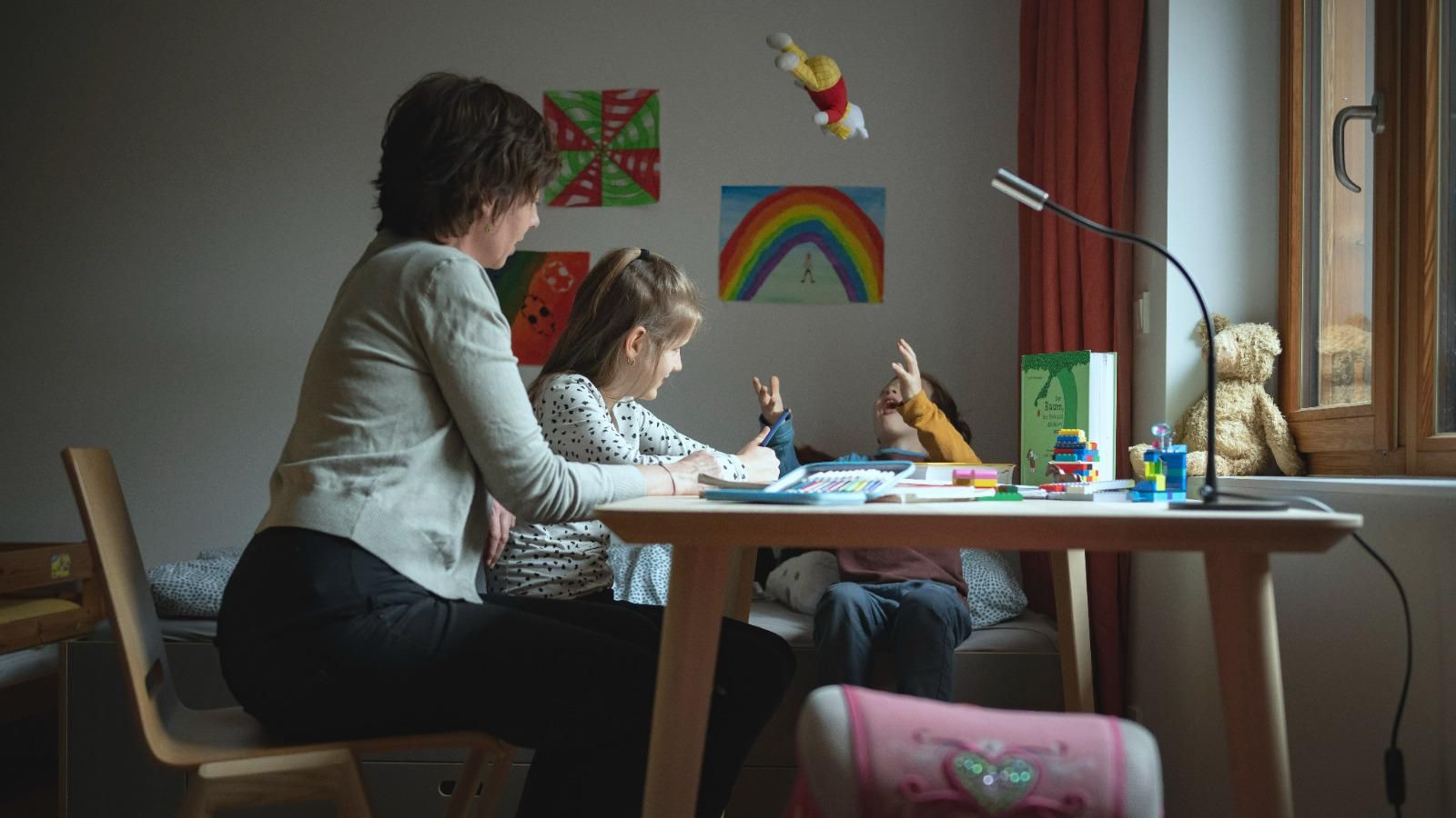 Sozialpädagogin lernt mit einem 10-jährigen Mädchen am Schreibtisch, im Hintergrund hängen selbstgemalte Bilder an der Wand