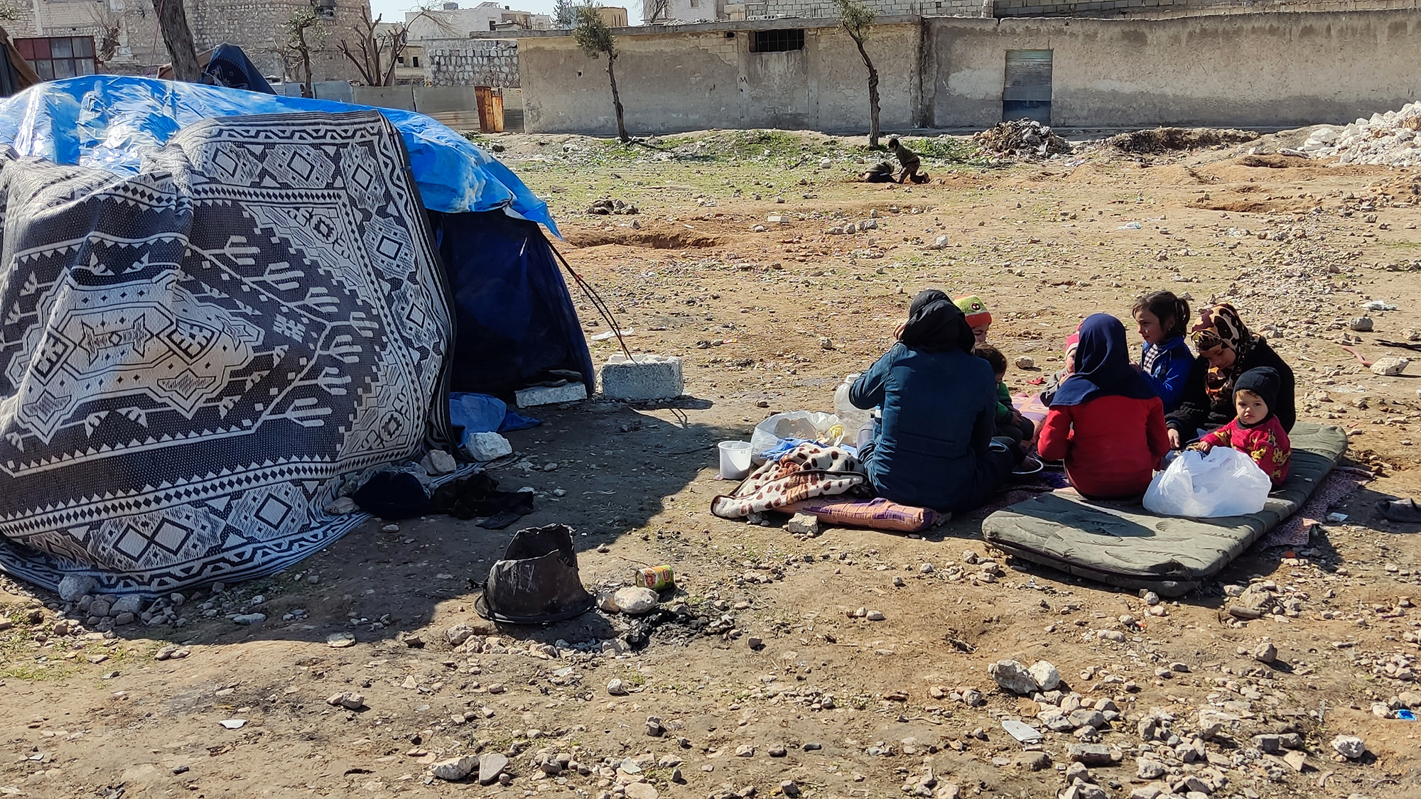 junge Menschen sitzen auf einer dünnen Matratze auf der Erde neben einem provisorisch aufgestellten Zelt