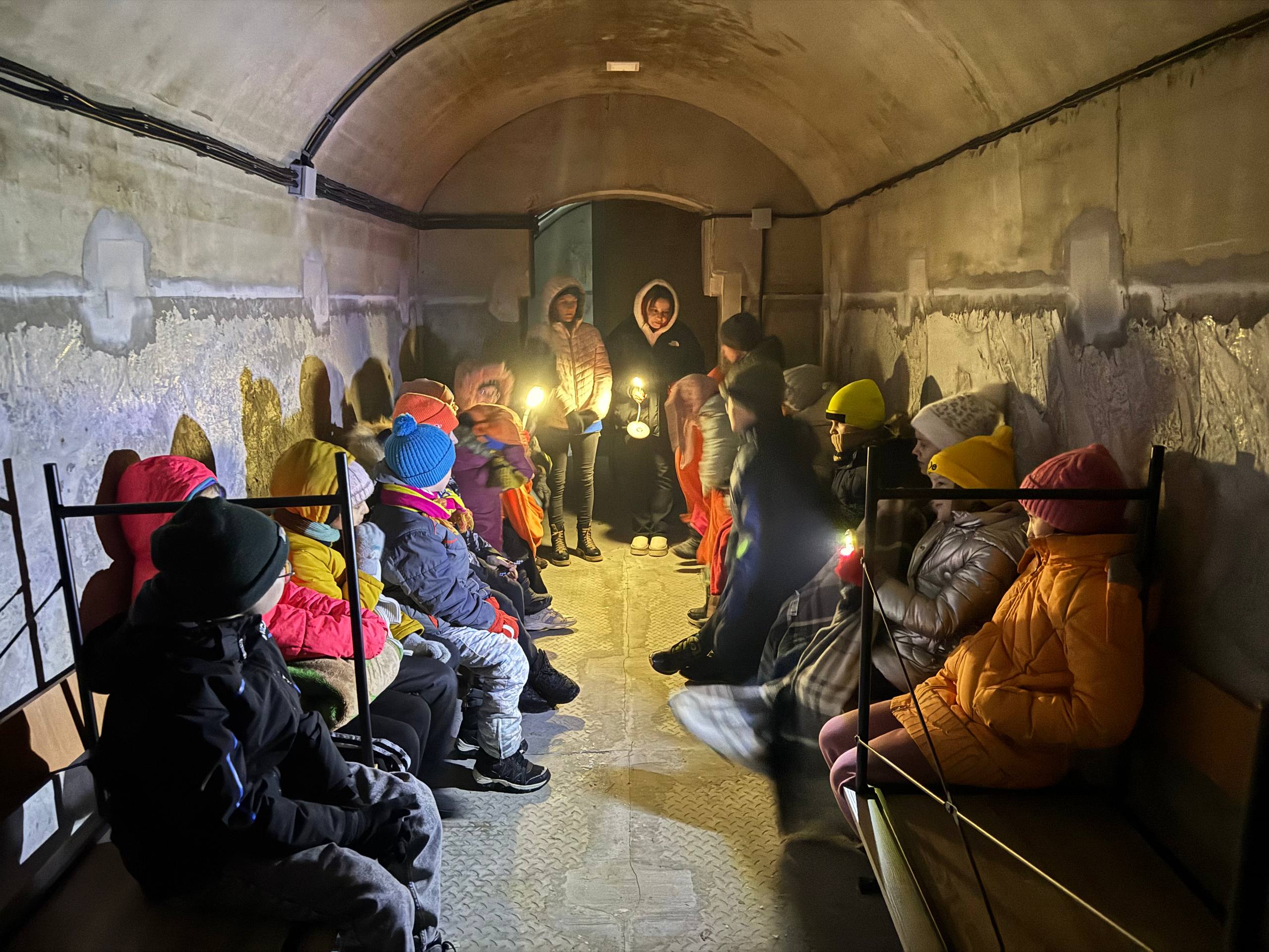 Kinder sitzen mit Mäntel und Kapuzen in einem Schutzraum.